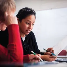 Two women in a classroom reviewing documents while a man is at a whiteboard in the background.